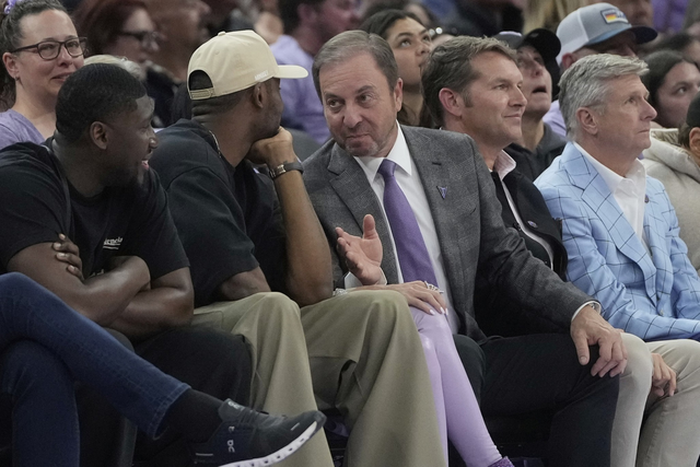 A group of men sit closely together in arena seats, engaged in conversation. One man in a suit is gesturing with his hand. The crowd is attentive and focused.